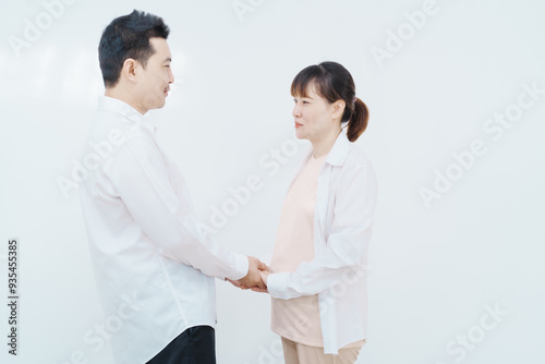 A happy Asian couple, expecting a baby, embraces in a cozy living room. The pregnant woman smiles warmly, while the man lovingly supports her, capturing a moment of joyful anticipation.