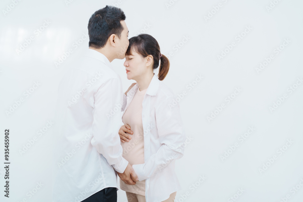 A happy Asian couple, expecting a baby, embraces in a cozy living room. The pregnant woman smiles warmly, while the man lovingly supports her, capturing a moment of joyful anticipation. A happy Asian couple, expecting a baby, embraces in a cozy living room. The pregnant woman smiles warmly, while the man lovingly supports her, capturing a moment of joyful anticipation.