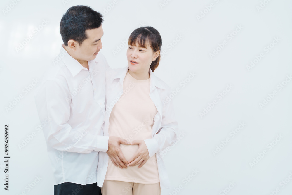 A happy Asian couple, expecting a baby, embraces in a cozy living room. The pregnant woman smiles warmly, while the man lovingly supports her, capturing a moment of joyful anticipation. A happy Asian couple, expecting a baby, embraces in a cozy living room. The pregnant woman smiles warmly, while the man lovingly supports her, capturing a moment of joyful anticipation.