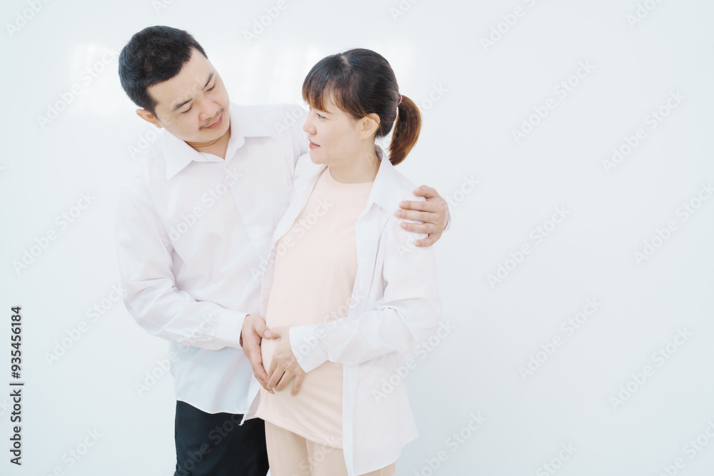A happy Asian couple, expecting a baby, embraces in a cozy living room. The pregnant woman smiles warmly, while the man lovingly supports her, capturing a moment of joyful anticipation. A happy Asian couple, expecting a baby, embraces in a cozy living room. The pregnant woman smiles warmly, while the man lovingly supports her, capturing a moment of joyful anticipation.