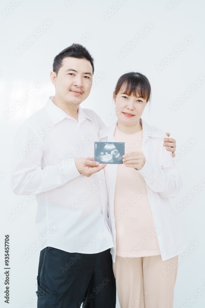 A happy Asian couple, expecting a baby, embraces in a cozy living room. The pregnant woman smiles warmly, while the man lovingly supports her, capturing a moment of joyful anticipation. A happy Asian couple, expecting a baby, embraces in a cozy living room. The pregnant woman smiles warmly, while the man lovingly supports her, capturing a moment of joyful anticipation.
