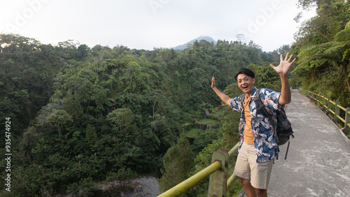 Happy young asian man wearing summer clothing and backpack is taking photo at Jembatan Plunyon, Kalikuning, Cangkringan, Sleman hills. Concept for solo traveling photo