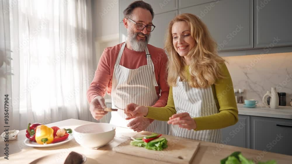 Beautiful Caucasian couple widely smiling while preparing food together. Happy people wearing aprons and cooking sandwiches for breakfast. Home routine. Man helping his wife in kitchen. Beautiful Caucasian couple widely smiling while preparing food together. Happy people wearing aprons and cooking sandwiches for breakfast. Home routine. Man helping his wife in kitchen.