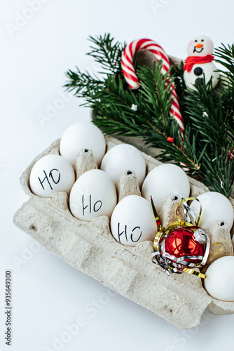 in the egg tray there are eggs with the inscription HO-ho-ho and a red Christmas tree ball in place of one egg, on a white background and fir branches and decorations