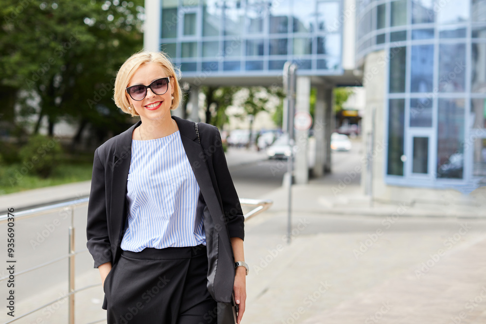 Fototapeta premium Beautiful blonde young woman wearing suit and walking on the street. A woman dressed in a business style walks around the city