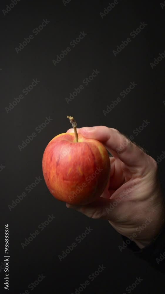 Hand holding a red apple between fingers, moving around, dark black background vertical video with copy space for text