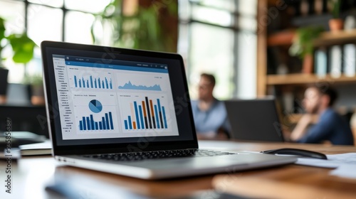 A well-organized modern workspace featuring a laptop on a wooden desk displaying financial metrics and graphs, with a background of professionals engaged in collaborative discussion. photo