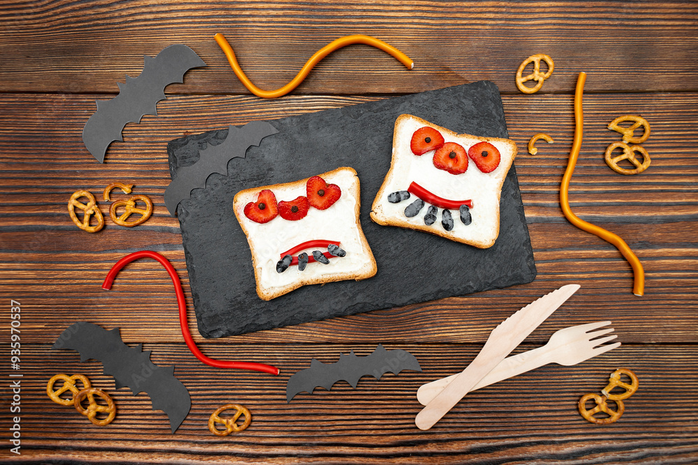 Obraz premium Funny monster face on halloween sandwich toast bread with butter,strawberry,sweets,cutting serving board,bats on black background.Kids child sweet dessert breakfast lunch food