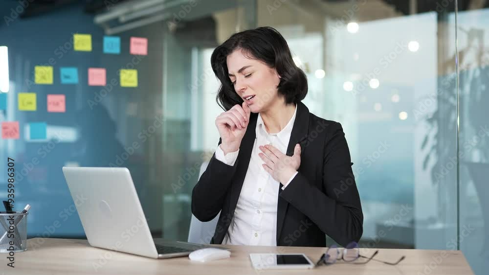 Female office worker suffering from cough and cold during work hours. Woman in professional attire holding chest sitting at desk at workplace in office, indicating discomfort from respiratory illness Female office worker suffering from cough and cold during work hours. Woman in professional attire holding chest sitting at desk at workplace in office, indicating discomfort from respiratory illness