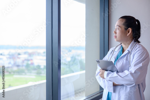 Asian female doctor contemplating looking through window standing at hospital building holding digital tablet