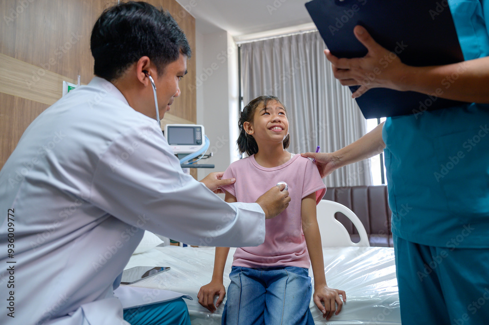 Obraz premium Asian male doctor using stethoscope to check pulse of girl in hospital examining and treating child patient with nurse beside him