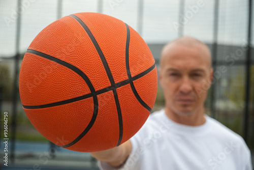 Portrait of sporty adult man holding orange basketball ball in hand on sports court. Focus on ball. Close up.