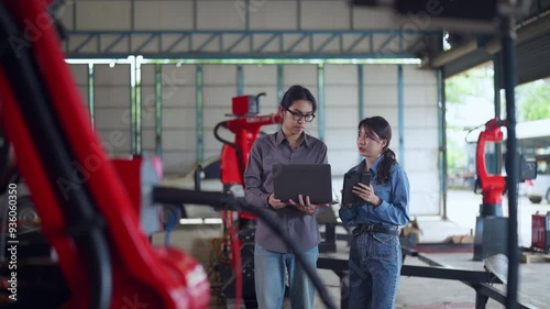 Two engineers collaborating in an industrial workshop, using a laptop and discussing work, surrounded by red robotic machinery.