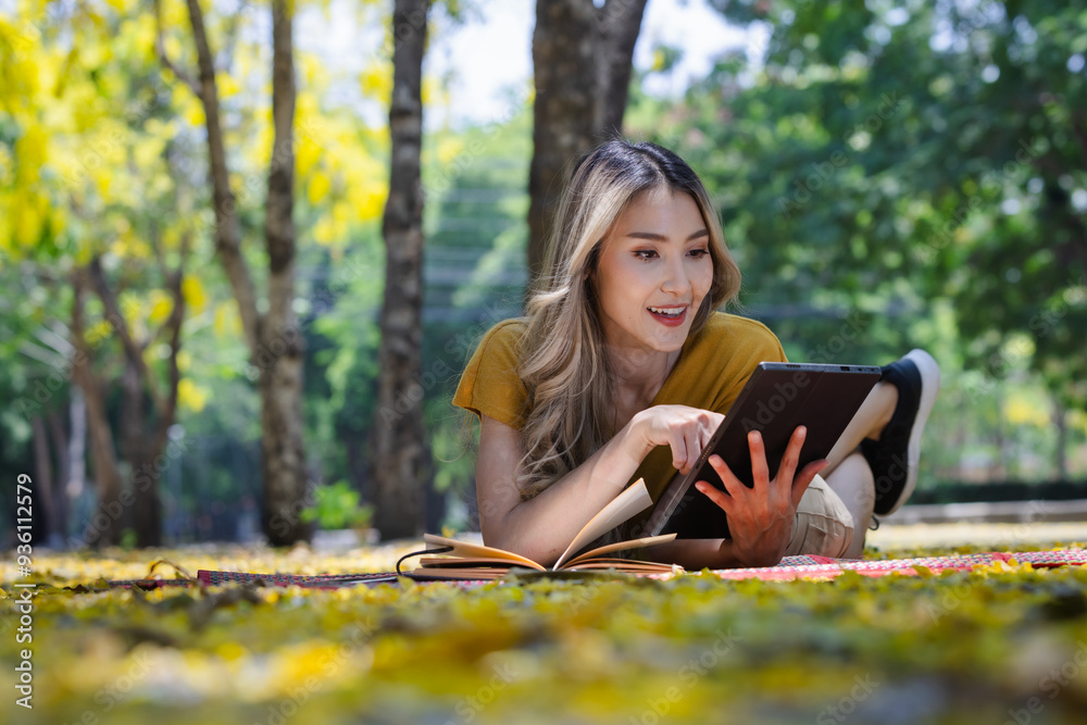 Smiling Woman Relaxing Outdoors on a Blanket, Reading a Book and Using a Tablet in a Sunlit Park with Yellow Leaves and Greenery