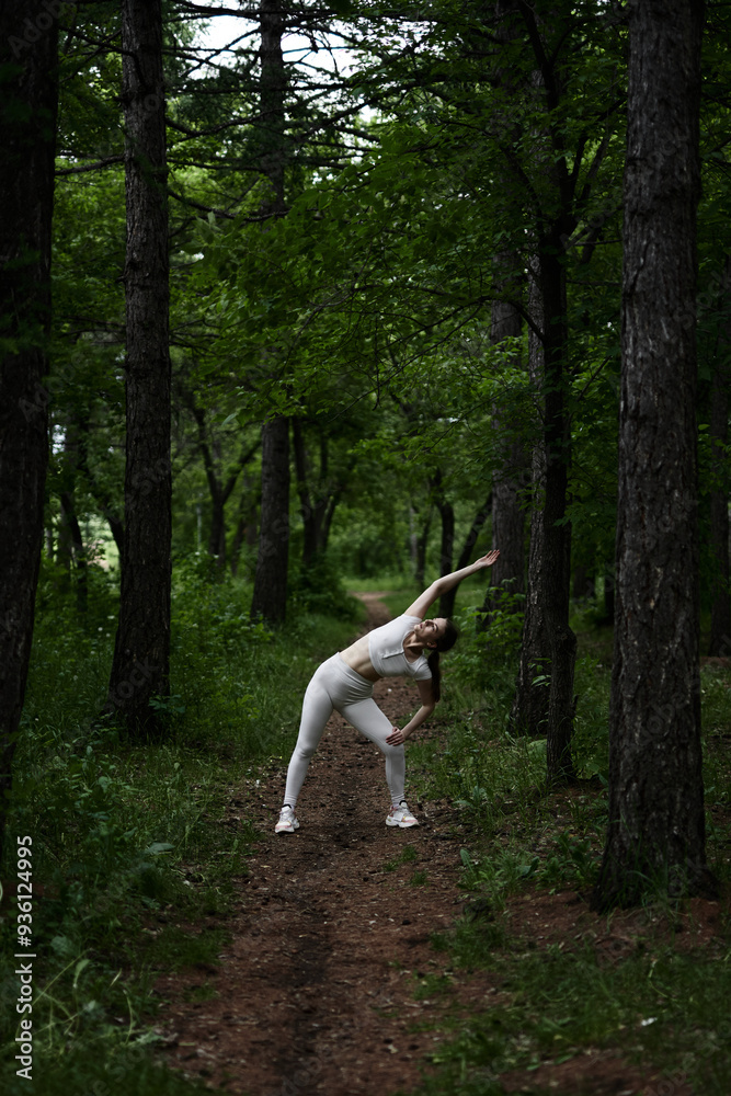 Fototapeta premium Young caucasian woman in white sportswear exercising in green forest park