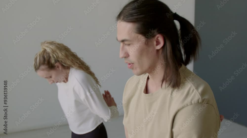 Young multiracial man leaning back, bending forward with hands behind his back and slowly rolling up while warming up with female dance partner before rehearsal