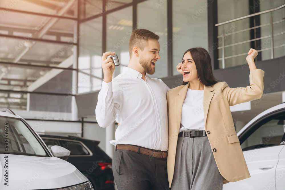 Happy young lovely couple in casual wear hugging while buying first new family car together in dealership. Man shows car keys in showroom Happy young lovely couple in casual wear hugging while buying first new family car together in dealership. Man shows car keys in showroom