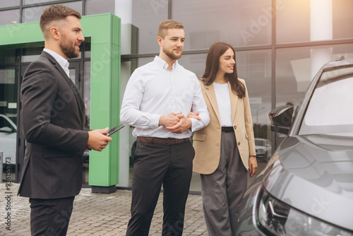 Young couple and vehicle manager standing outside auto showroom near car. Man and woman talking with sales manager.