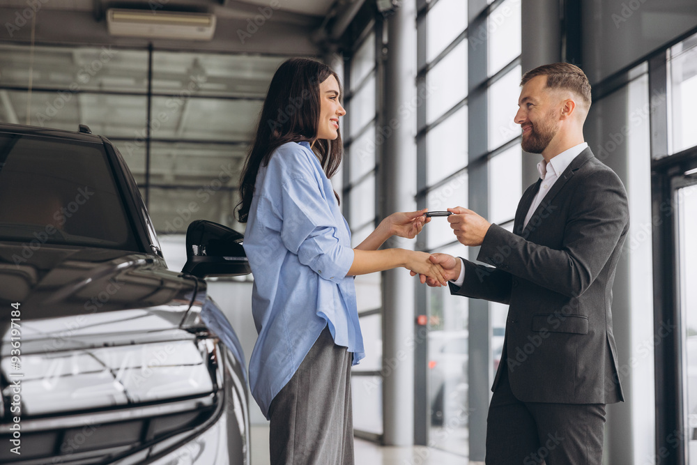 Smiling salesman or manager giving car keys and shakes hand to modern happy woman new car owner in car dealership