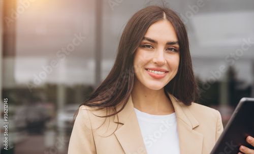 Close-up portrait of confident successful saleswoman in suit with digital tablet in hands standing in outdoor car dealership