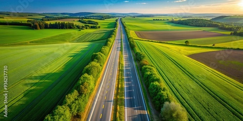 Aerial view of a straight, two-lane asphalt highway cutting through a vast, lush agricultural landscape of green fields and rolling hills in rural countryside. photo