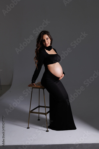 A beautiful pregnant woman with long dark hair with styling and makeup. A brunette waiting for a baby, sit on high chair in a long black dress. Grey background in the studio, fan.