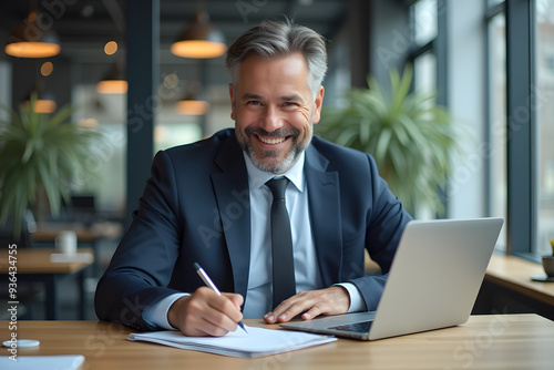 Happy middle aged professional business man company executive ceo manager wearing suit sitting at desk in modern office working on laptop computer and writing notes, portrait at workplace.