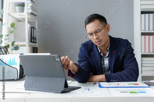 Businessman is frowning and reading bad news on a tablet screen while sitting at his desk in the office