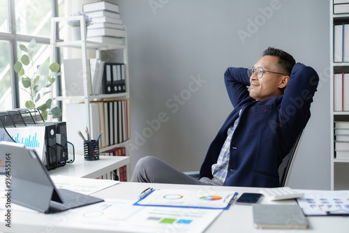 Asian businessman relaxing with his hands behind head sitting at his desk in a luminous office photo