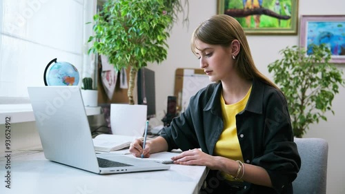 Teenage female studying at home, sitting at desk with laptop computer, writing in notebook