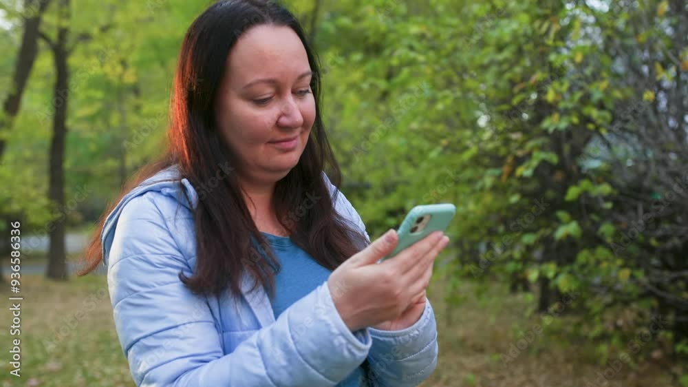 Smiling woman using smartphone while standing against trees. Beautiful female is socializing online at park. She is wearing jacket while spending leisure time during winter.