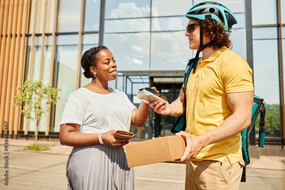 Obraz premium Smiling recipient receiving package from delivery person outside modern building. Both individuals are engaging in friendly interaction while person in helmet hands over the item