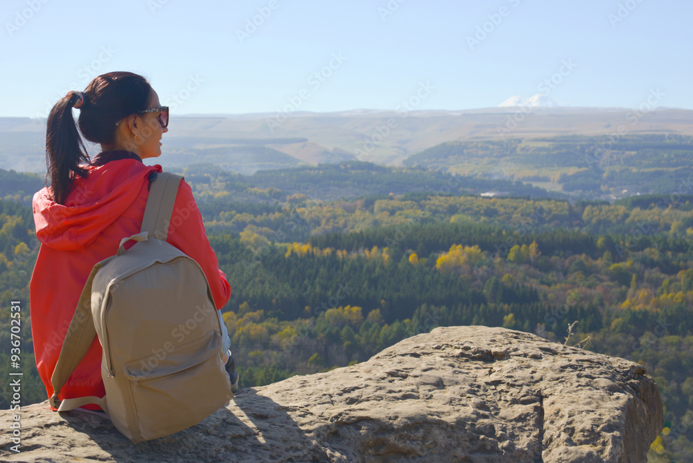 A girl tourist with a backpack sits on the top of a mountain and enjoys the beautiful views. A walking woman in an orange jacket rests on a rock, looking at the beautiful sunny landscape of the