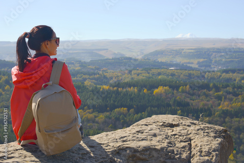 A girl tourist with a backpack sits on the top of a mountain and enjoys the beautiful views. A walking woman in an orange jacket rests on a rock, looking at the beautiful sunny landscape of the