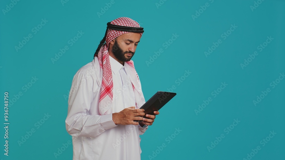 Young adult navigating on gadget in studio, browsing websites online on modern device. Muslim guy in traditional costume using tablet to check internet pages, chatting on social media. Young adult navigating on gadget in studio, browsing websites online on modern device. Muslim guy in traditional costume using tablet to check internet pages, chatting on social media.