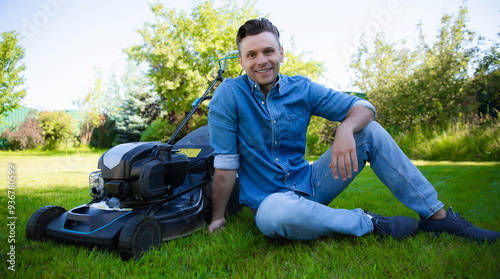 A smiling man in a denim shirt sits on a well-manicured lawn next to a lawn mower, enjoying a moment of rest after yard work. Satisfaction, outdoor relaxation, and the reward of a job well done.