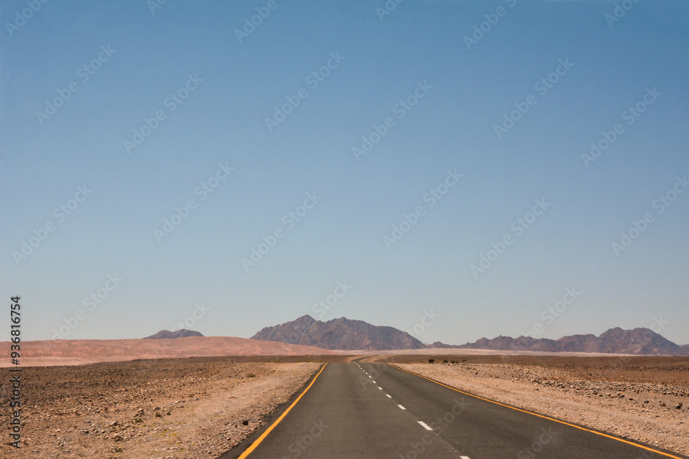 A flat road in the desert in perspective leading to the mountains under the blue sky A flat road in the desert in perspective leading to the mountains under the blue sky