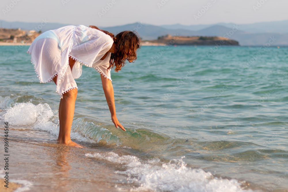 Naklejka premium A young girl in white dress touching the water on the beach