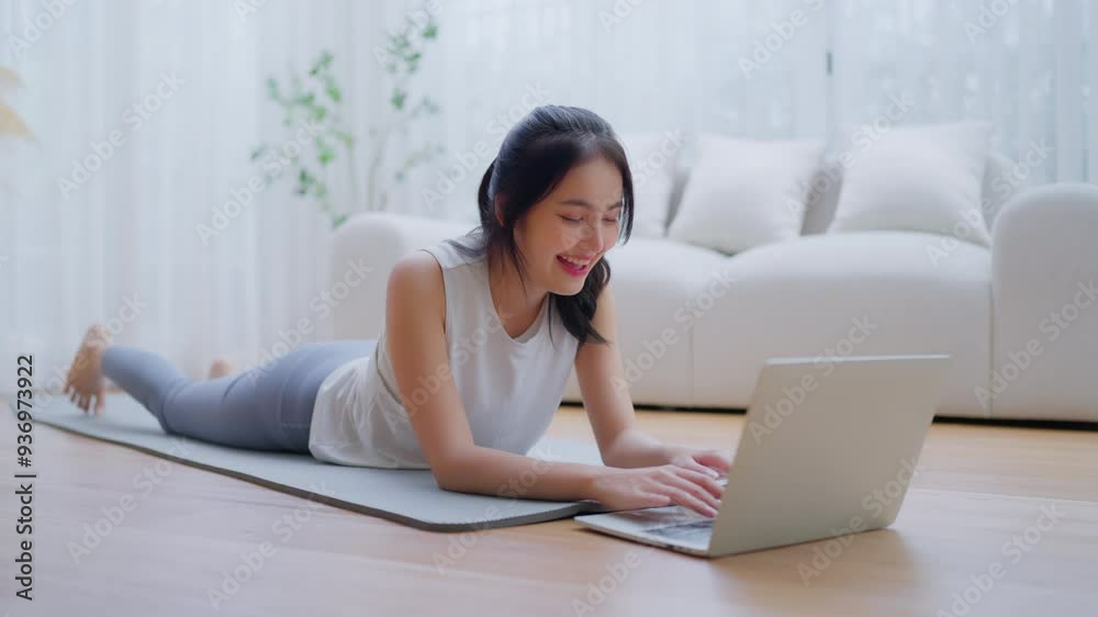 Young Asian woman lying on the floor on yoga mat in front of laptop and training at home. Beautiful smiling woman doing stretching exercise while communicates with a trainer online before workout Young Asian woman lying on the floor on yoga mat in front of laptop and training at home. Beautiful smiling woman doing stretching exercise while communicates with a trainer online before workout