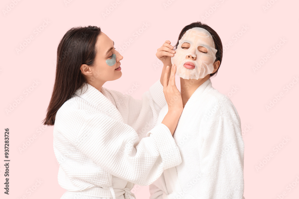 Young women in bathrobes making cosmetic procedure to each other on pink background Young women in bathrobes making cosmetic procedure to each other on pink background