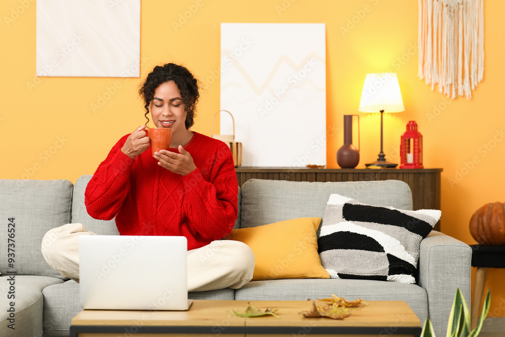 Beautiful young happy African-American woman in stylish outfit with laptop, cup of tea and autumn leaves sitting on sofa at home Beautiful young happy African-American woman in stylish outfit with laptop, cup of tea and autumn leaves sitting on sofa at home