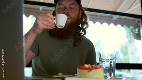 Contented curly young man drinking aromatic espresso from small cup near cheesecake open laptop and glass of water, low angle. photo