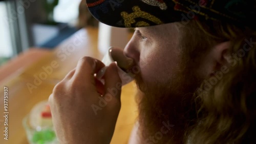 Side view of thoughtful red-bearded young guy drinking espresso from small mug sitting at wooden table in coffee shop looking out window to street. photo