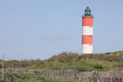 Vue extérieure ensoleillée du phare de Berck dans le Pas-de-Calais en France. Structure cylindrique peinte en rouge et blanc. Dunes et oyats verts au premier plan. Ciel bleu en arrière plan.