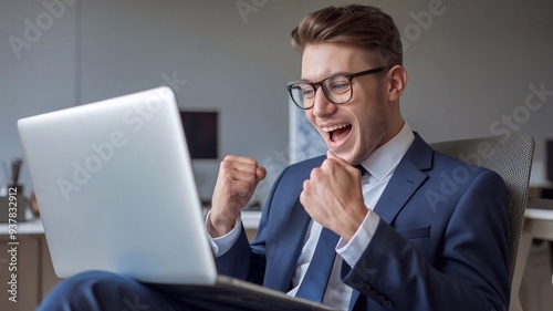 Excited young businessman sitting in his office, looking at the laptop computer device with surprised reaction. Wearing glasses andblue suit. Male person or entrepreneur shocked, wow face expression photo