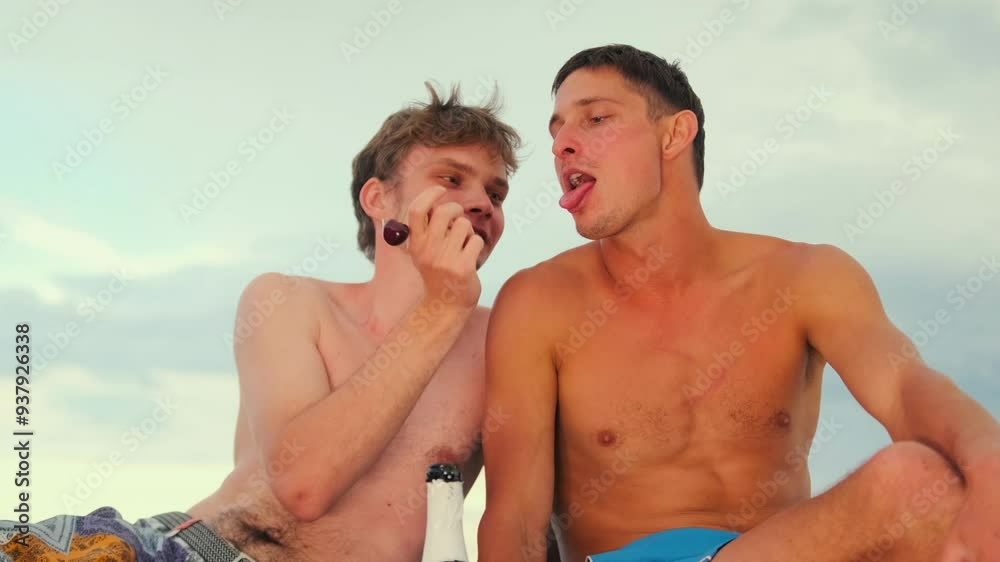 Two guys frolicking on a romantic date or doing cherries feeding each other from their hands against a cloudy sky sitting shirtless on the sand. Two guys frolicking on a romantic date or doing cherries feeding each other from their hands against a cloudy sky sitting shirtless on the sand.