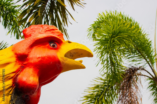 Close-up head of colorful ceramic rooster figurine against blurred palm leaves and sky in Ayutthaya, Thailand. A popular religious offering in Thai cultures photo