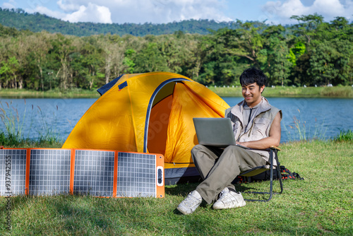 Young Asian man working remotely with laptop at a scenic campsite using portable solar panels for sustainable energy in nature