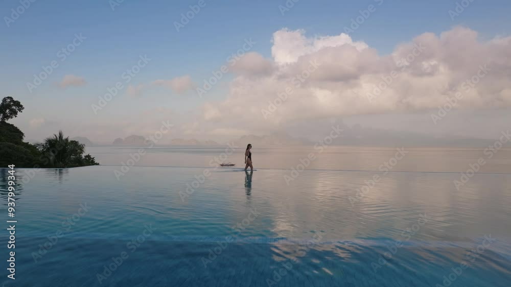 Aerial view of sexy woman in swimwear relaxing in luxury infinity pool, looking out over the ocean on exotic summer vacation at a spa hotel with beautiful sky clouds reflected in the water at sunrise Aerial view of sexy woman in swimwear relaxing in luxury infinity pool, looking out over the ocean on exotic summer vacation at a spa hotel with beautiful sky clouds reflected in the water at sunrise