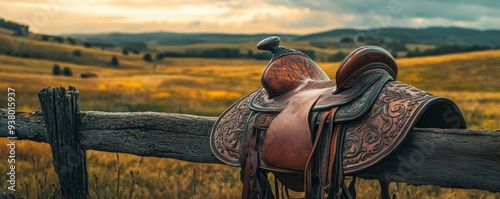 Beautiful, tooled leather western saddle is sitting on a rustic, wooden fence with rolling hills and the warm glow of sunset in the background